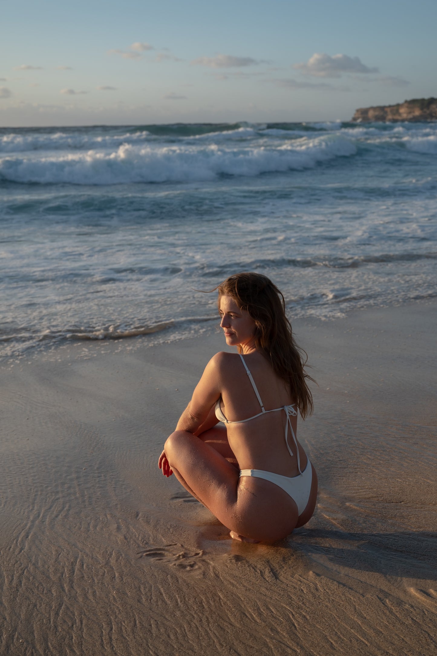 A woman at a beach wearing a white triangle top with adjustable straps, tie back and silver detailing .