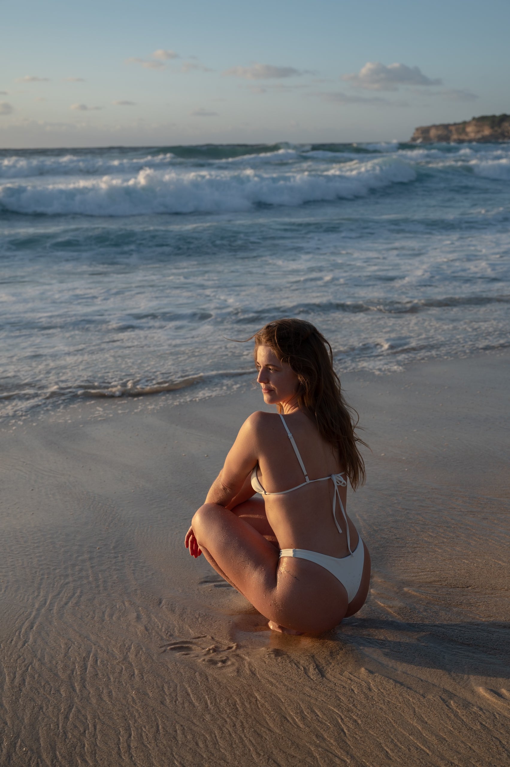 A woman at a beach wearing a white triangle top with adjustable straps, tie back and silver detailing .