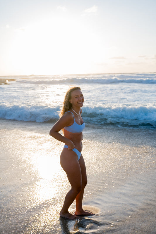 A woman standing on a beach at sunset wearing a blue swimwear top thats scoop neck and the matching bottoms.