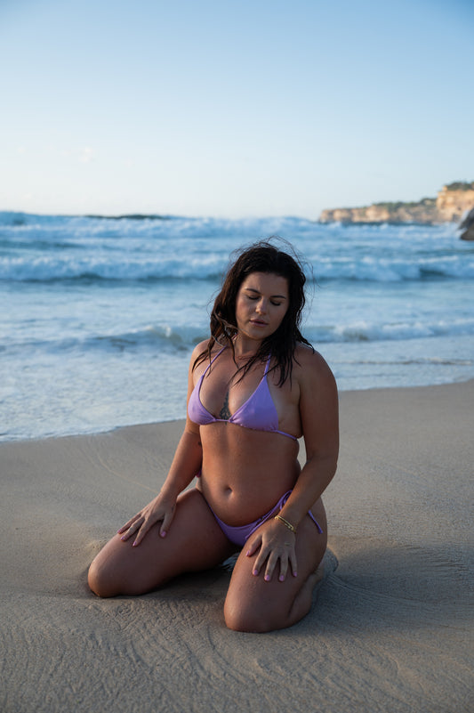 A woman sitting on a sandy beach wearing a lavender-colored triangle bathing suit top with ties, matching with purple tie bottom. Front View.